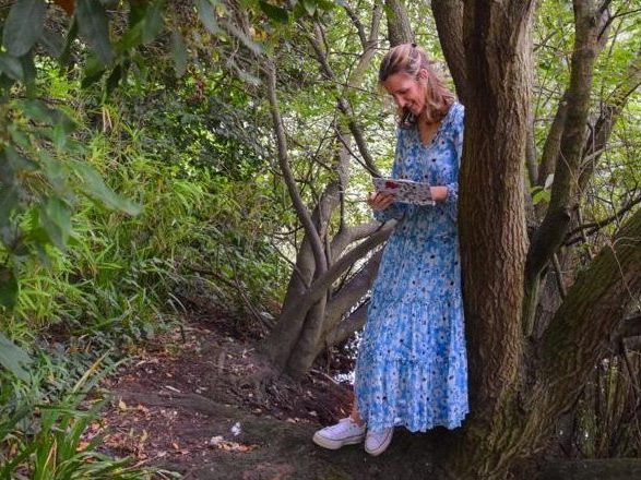 Author and Journalist Jemimah Wright leans against a tree, writing in her journal, wearing a blue dress.